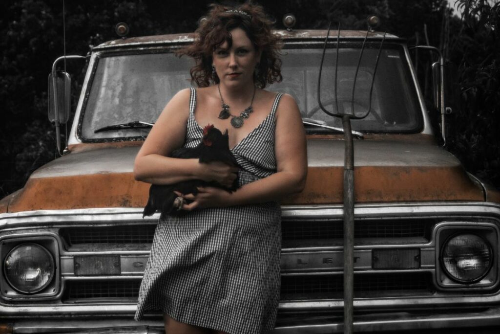 First Friday Artist Taylor Endres poses, leaning on an old car, chicken in hand and pitchfork leaning beside.